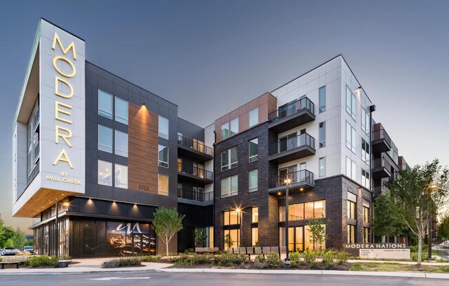 Modera Nations features bold architecture, mixed materials, and illuminated signage anchoring the neighborhood streetscape at dusk.