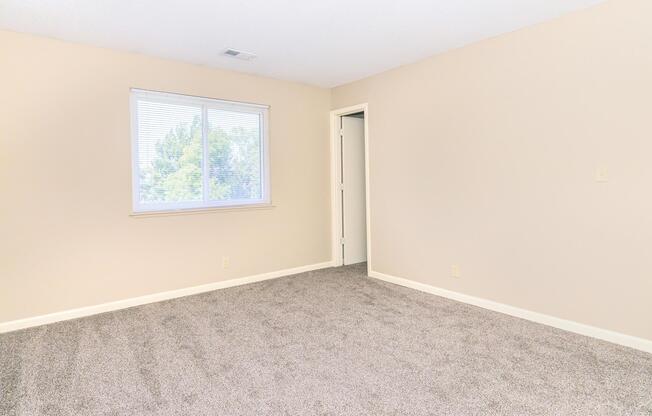 A neutral-colored bedroom with beige walls and light gray carpet. There's a single window with white blinds letting in natural light. A closed door is visible on the right, contributing to a spacious, empty feel. The room is unfurnished, making it suitable for personal customization.