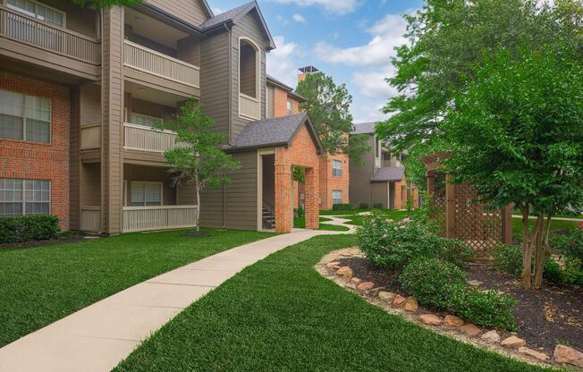 A well-maintained apartment complex with three buildings, surrounded by lush green grass and neatly trimmed shrubs. The pathway is paved, leading through the landscaped area with a stone border, creating an inviting atmosphere in a residential setting. The sky is partly cloudy, enhancing the peaceful environment.