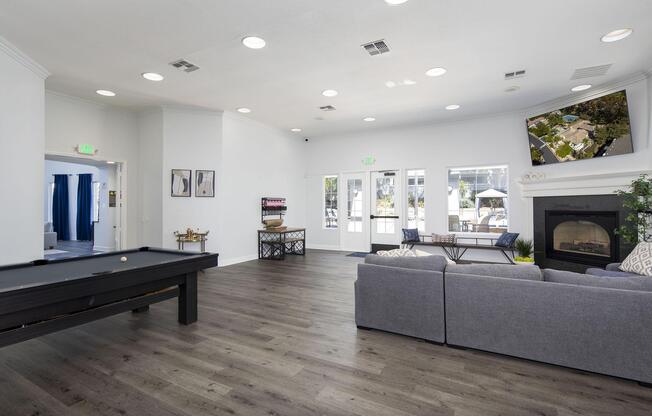 A spacious lounge area featuring a grey couch, a pool table, and a modern fireplace. Large windows allow natural light, while a TV is mounted on the wall, showing an outdoor view. The decor is contemporary, with light-colored walls and wooden flooring, creating an inviting atmosphere for relaxation and socializing.