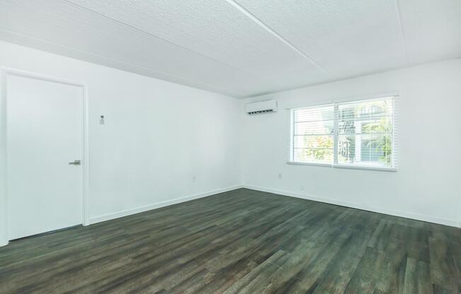 A bright, empty room with white walls and a textured ceiling. It features a large window with white blinds and a small air conditioning unit mounted above the window. The floor is covered in dark laminate, creating a modern and clean aesthetic. A closed door is visible on the left side of the image.