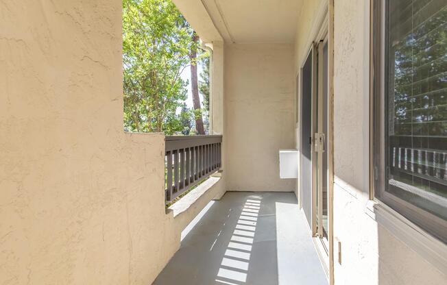 A small balcony with a light-colored surface, bordered by a low railing. Sunlight casts shadows from the railing onto the floor, and there are trees visible in the background, creating a serene outdoor space. The balcony is empty and offers a view of the surrounding greenery.