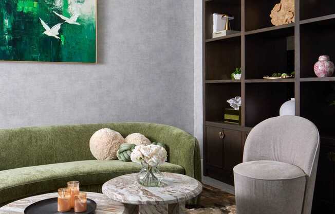A living room with a green sofa, a round table with candles, a grey armchair, and a shelf with various decorative items.
