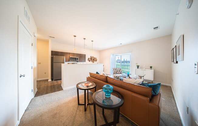 Living room looking into kitchen, light tan carpet with brown leather couch, end tables behind couch, photos on the wall to the right, kitchen enternce on the left, tan walls.