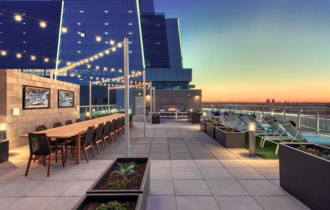 A rooftop patio with a long table and chairs, string lights, and a view of the city skyline at dusk.