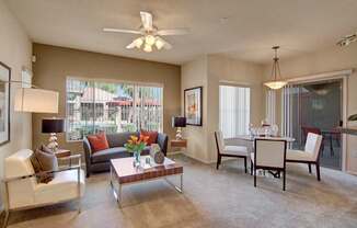 A spacious living and dining area here at Dobson Towne Center featuring plush carpet, neutral walls, and a ceiling fan with light. A cozy sofa and accent chair surround a modern coffee table, while a dining space sits beside large windows and a sliding glass door that provides natural light and access to a private patio with community views.