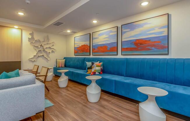 A modern lounge area featuring a blue upholstered bench with colorful pillows, two light-colored side tables, and a grey sofa. The walls are adorned with abstract paintings in vibrant colors, and there is a textured wall art piece. The flooring is wooden, creating a cozy atmosphere.