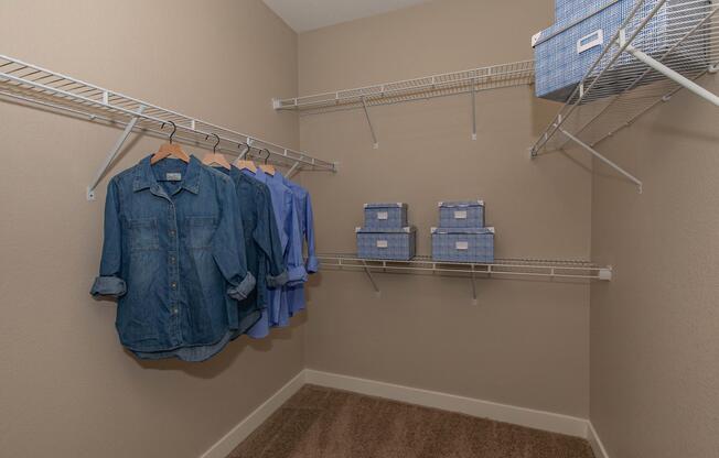 A well-organized closet featuring two hanging denim shirts and a light blue blouse on hangers. Two shelves are lined with blue patterned storage boxes. The interior walls are painted in a beige color, and the floor is covered with a soft carpet.