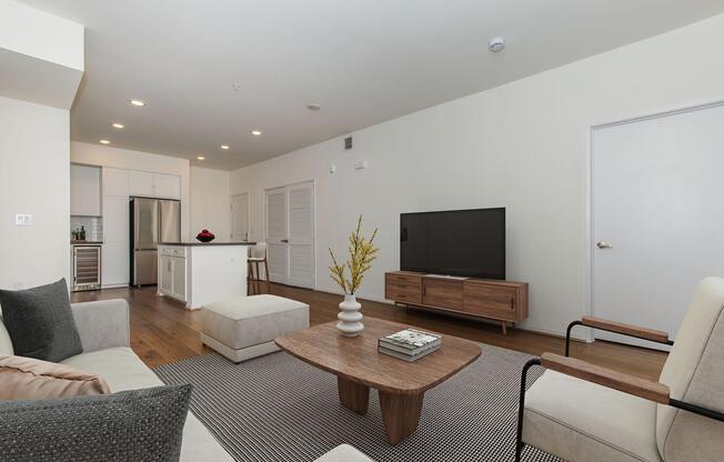 A modern living room featuring a light-colored sofa, a wooden coffee table, and a minimalist entertainment center with a large TV. The space has light walls, wooden floors, and a stylish area rug. A kitchen is visible in the background, showcasing contemporary appliances and cabinetry.