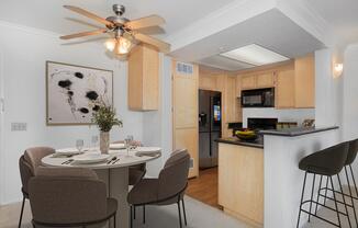 A modern kitchen and dining area featuring a round table set for four, elegant dining chairs, a ceiling fan, and wooden cabinetry. The kitchen includes a black countertop, stainless steel appliances, and a framed abstract art piece on the wall. Natural light brightens the space.
