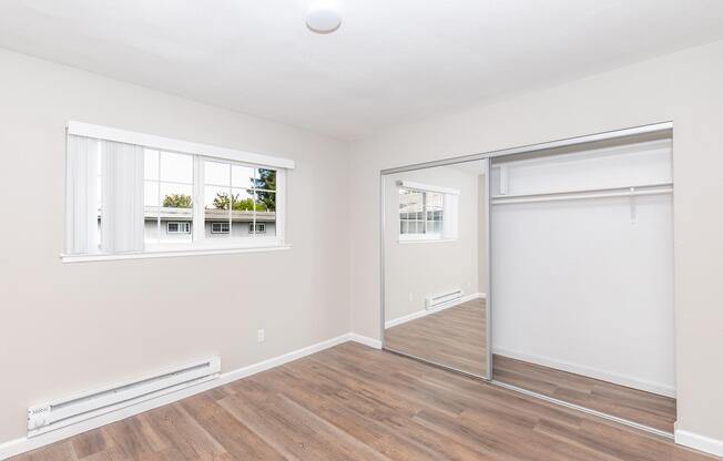 A well-lit, empty room featuring a large window with white blinds and a mirrored closet. The walls are painted a soft beige, and the flooring is modern wood laminate. The room is designed with simplicity and spaciousness in mind, suitable for various furnishings.