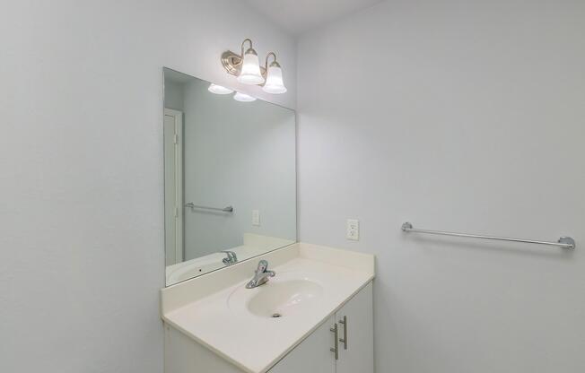A modern bathroom setting featuring a white countertop with a single sink, a large mirror above it, and light fixtures. The walls are painted a soft color, and there is a towel bar mounted on the wall. The overall look is clean and minimalist.