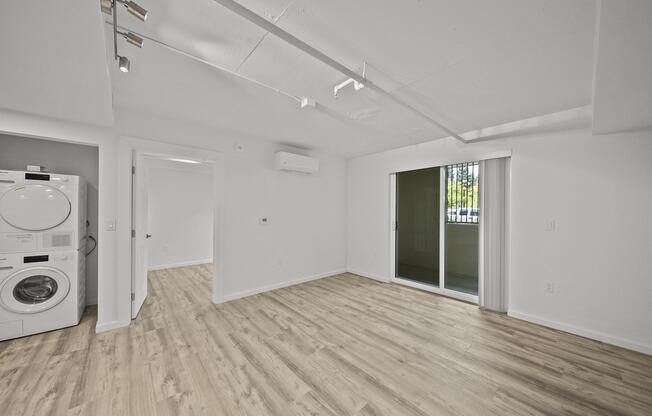 A bright, modern room with light wood flooring, featuring a wall-mounted air conditioner and a sliding glass door leading to an outdoor area. On the left, there is a stacked washer and dryer unit. The walls are painted white, contributing to a clean and spacious atmosphere.