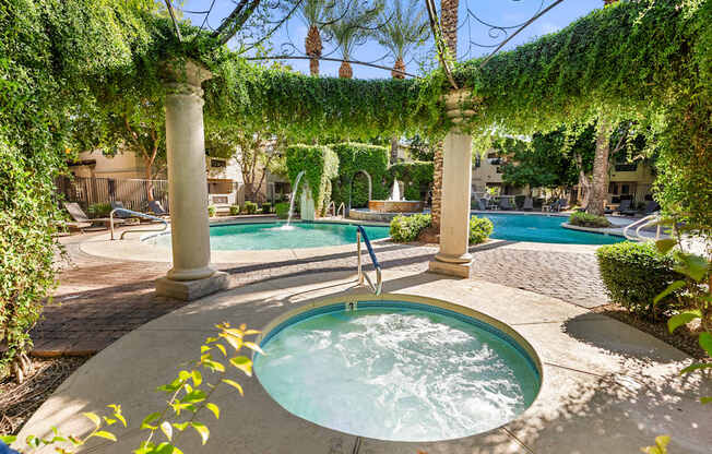 A hot tub sits in the middle of a patio under a vine covered pergola.