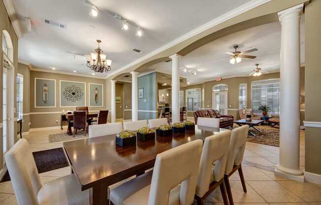 A spacious clubhouse interior here at Mission Green featuring a large dining table with upholstered chairs, decorative columns and archways, elegant chandeliers and ceiling fans, tiled flooring, and adjacent lounge seating with sofas and coffee tables, creating a welcoming shared space for dining, relaxing, and gathering with neighbors.