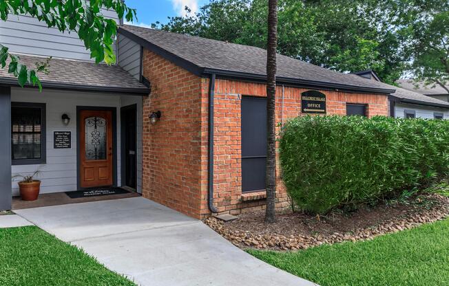 A brick building with a sloped roof and a wooden front door. The entrance features a small porch with a welcome mat, and there are potted plants on either side. Lush green grass and hedges surround the pathway leading to the door. A sign labeled "Residential Office" is mounted on the building's exterior.