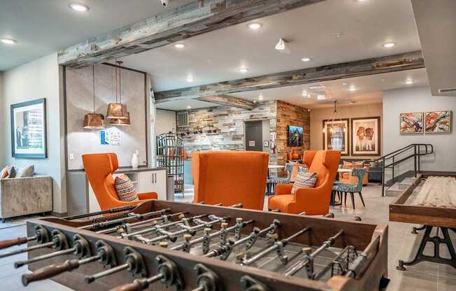 A foosball table is in the middle of a room with orange chairs and a stone wall.