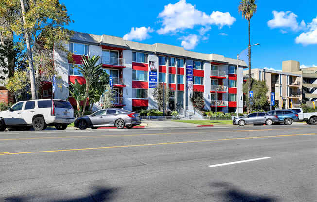 A multi-story building with a red, white, and blue color scheme is surrounded by parked cars and palm trees.