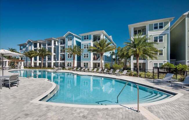 A swimming pool surrounded by lounge chairs and palm trees in front of apartment buildings.