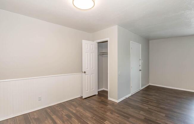 A well-lit, empty room with light gray walls and wooden flooring. There is a closed white door and a closet with an open door on the left side. The room features a simple ceiling light fixture and a small section of wainscoting near the floor.