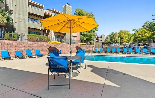 A yellow umbrella shades a table with blue chairs by a pool.
