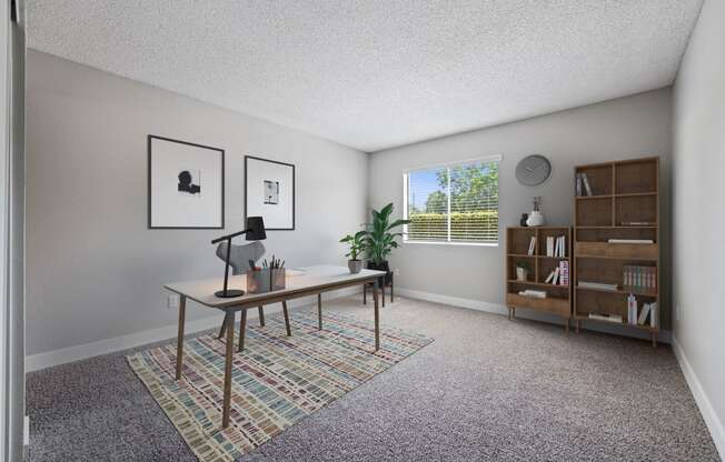 Apartments In Colton, CA - Ardella At Reche Canyon - Home Office With Carpet Flooring, Spacious Desk With Lamp, Chair, Potted Plants, Bookshelves, And Window With Blinds.