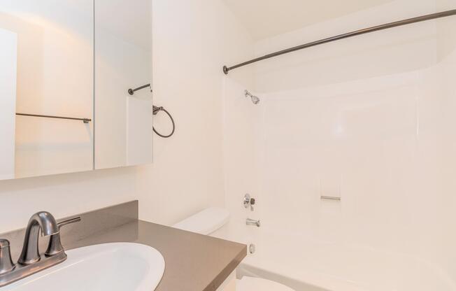 A clean, modern bathroom featuring a white bathtub and shower combination, a sleek black countertop with a sink, and a mirror above the sink. The walls are plain and bright, enhancing the spacious feel, with a black towel bar and minimalistic fixtures. The overall design is simple and contemporary.