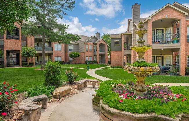 A serene courtyard featuring a decorative fountain surrounded by colorful flowers and neatly landscaped greenery. In the background, brick apartment buildings are visible under a partly cloudy sky, creating a peaceful residential atmosphere. Pathways wind through the garden, inviting residents to stroll.