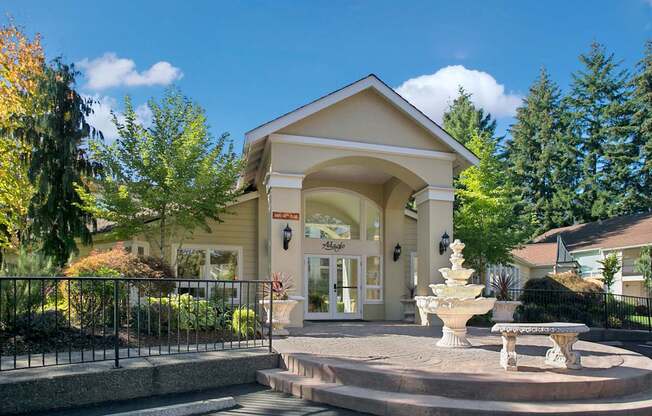 A grand clubhouse entrance here at Adagio featuring an arched covered entryway, tall columns, and glass doors accented by decorative lighting, with a classic tiered fountain and stone seating in the foreground, while manicured landscaping, mature trees, and wrought-iron railings frame a welcoming focal point for the community.