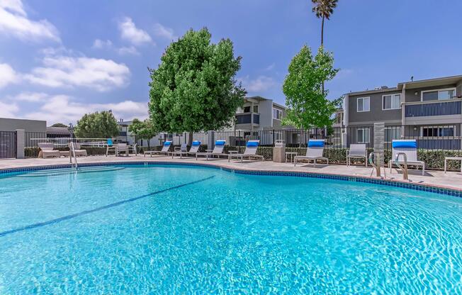 A clear blue swimming pool surrounded by lounge chairs, with green trees and modern apartment buildings in the background under a partly cloudy sky.