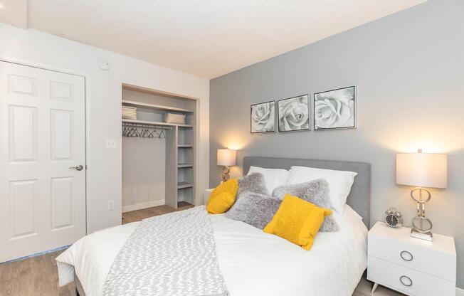 Cozy bedroom featuring a queen-sized bed with gray and white bedding, adorned with yellow pillows. A nightstand with a lamp sits beside the bed. The room has gray walls and a closet space with shelving. Three rose-themed pictures are hung above the bed, enhancing the decor.
