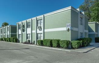 A green and white two-story building with a grid-patterned facade, featuring multiple windows and air conditioning units. There are neatly trimmed hedges along the sidewalk, and the sky is clear and blue, suggesting a bright day.
