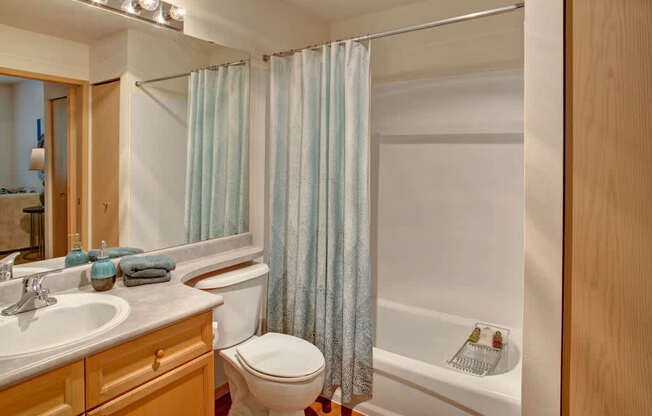 A clean bathroom here at Portsmith featuring a light wood vanity with a smooth countertop and integrated sink, a large wall mirror with bright overhead lighting, a toilet beside a tub and shower combination with a patterned shower curtain, and simple decorative accents that create a fresh, functional space.