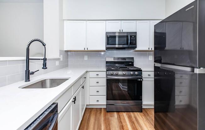 Modern kitchen with white cabinetry, stainless steel appliances including a microwave and a gas stove. A sleek black refrigerator is visible, and a contemporary sink with a black faucet is present. The flooring is wood, and the walls are painted in neutral tones, creating a clean and inviting atmosphere.