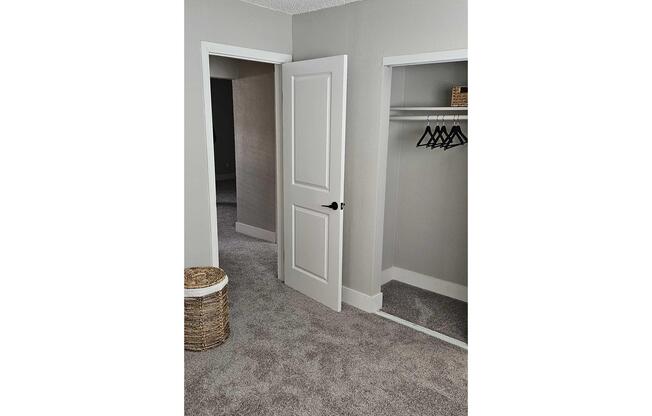 A minimalistic room featuring two doorways: one leading to a closet with a hanging rod and black hangers, and the other doorway appears to lead to another room. The walls are painted light gray, and the floor is covered in soft, beige carpet. A woven basket sits in the corner.