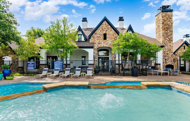 Pool with water feature in front of stone-accented clubhouse and poolside lounge chairs.