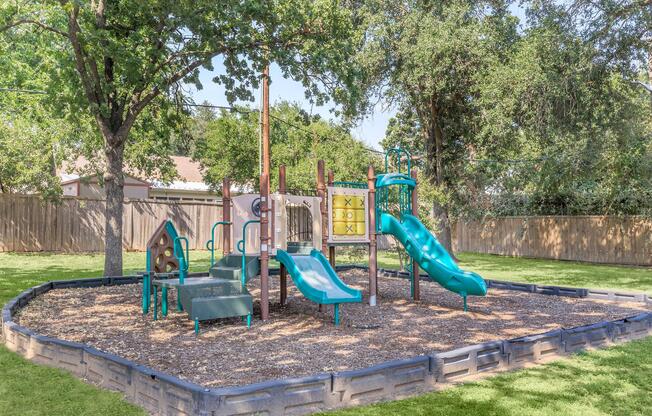 A playground featuring two slides, climbing structures, and swings, surrounded by grass and trees. The play area is bordered by a low wall and contains mulch on the ground for safety. Ideal for children to play and socialize.