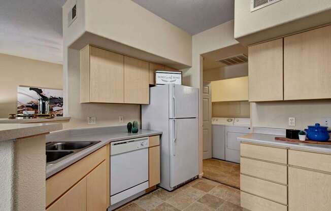 A functional kitchen here at Sonoma Landing showcasing light wood cabinetry, a full-size refrigerator, dishwasher, and double-basin sink set into neutral countertops, with tile flooring underfoot and an open layout that provides a clear view into an in-home laundry area with washer and dryer, creating a practical and connected living space.