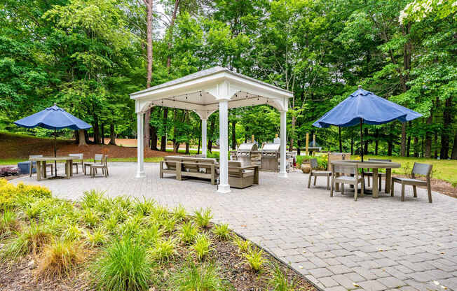A gazebo with a white roof is surrounded by benches and umbrellas in a park.