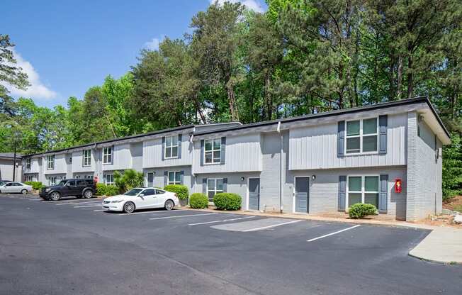 A parking lot in front of a grey building with trees in the background.
