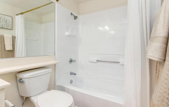 A clean, modern bathroom featuring a white bathtub with a shower curtain, a toilet, and a large mirror above a countertop. The walls are tiled in white, and there are two plush towels hanging on the right. A painting or decorative item is mounted on the wall.