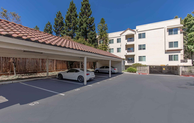 A parking lot with covered spaces and two parked cars, set against a backdrop of a four-story residential building with a red-tiled roof and surrounding trees. The lot is well-maintained, and the sky is clear, indicating a sunny day.