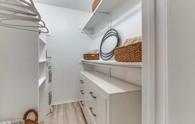 Fusion Fort Worth apartment walk in closet with wicker baskets on built in shelving, hangers on curtain rods, and white midcentury modern dresser.