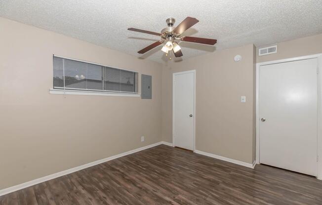 A spacious room featuring a light brown ceiling and wall color, a ceiling fan with light fixtures, laminate flooring, a small window with blinds, and two doorsâone leading to another room and one for a closet. The lighting is bright, highlighting the clean and simple design of the space.