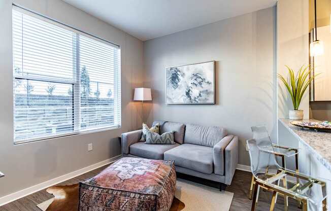 A living room with a grey couch, a patterned ottoman, and a painting on the wall.