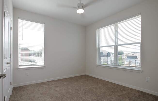A room with a carpet floor, a ceiling fan, and two windows with blinds.