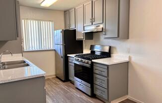 A modern kitchen featuring gray cabinets, a stainless steel stove and hood, a black refrigerator, and a double sink. The countertops are light-colored and there is a window with blinds, allowing natural light into the space. The flooring is wood-like laminate, giving a clean and contemporary look.