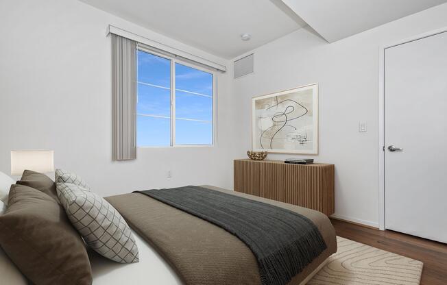 A modern bedroom featuring a bed with gray and white bedding, two decorative pillows, a wooden side table with a lamp, and a piece of abstract art on the wall. A window allows natural light to flood the space, and a wooden dresser is positioned against the wall. The room has a minimalist design with neutral colors and a clean aesthetic.