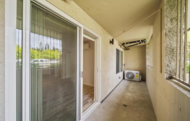 A view of a small covered balcony or porch area with a sliding glass door, concrete floor, and an air conditioning unit. The walls are beige, and there are large windows allowing natural light. The space appears to be part of an apartment or condo.