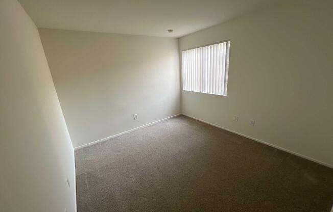 An empty room with light-colored walls and carpeted flooring. A window with vertical blinds allows natural light to enter, creating a bright atmosphere. The space is free of furniture, showcasing the room's dimensions and potential for use.